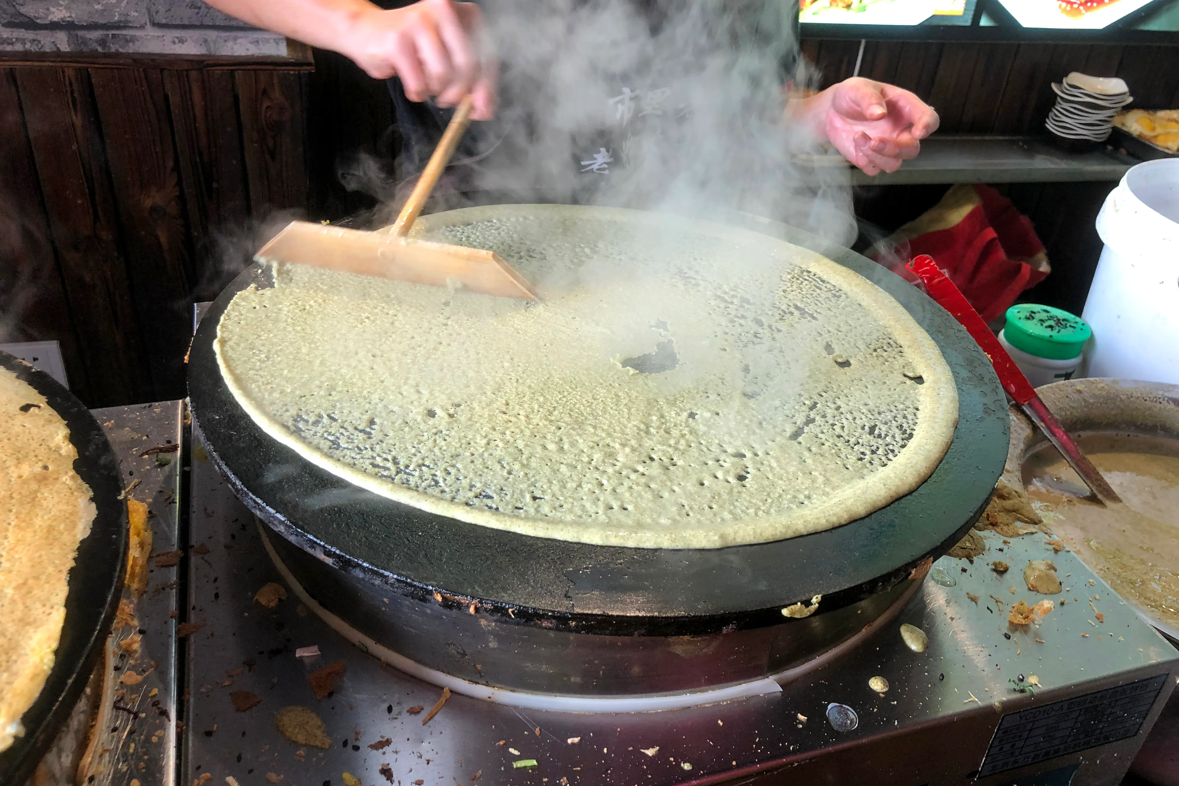 Street vendor making jianbing on a circular griddle — spreading batter and cracking an egg